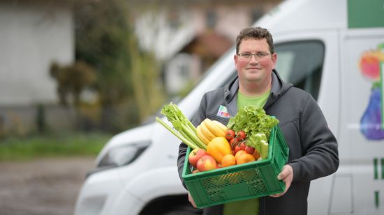 KI generiert: Ein Mann hält eine grüne Kiste voller Obst und Gemüse in den Händen. Im Hintergrund steht ein Lieferwagen.