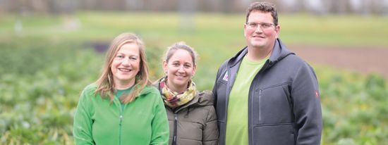 KI generiert: Das Bild zeigt drei Personen, die in legerer Kleidung vor einem grünen Feld stehen. Sie lächeln in die Kamera, und im Hintergrund ist landwirtschaftliche Natur zu sehen.