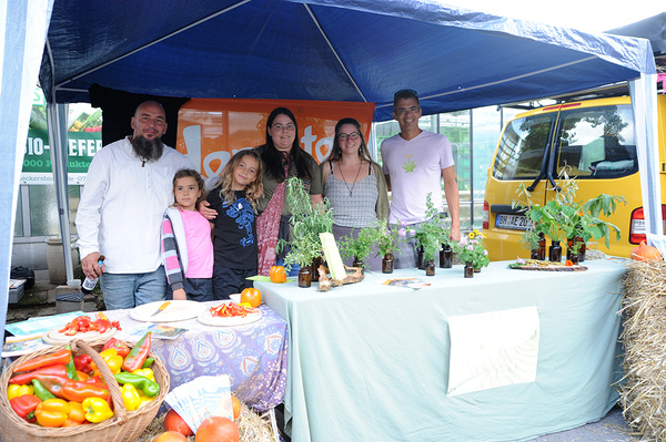 Ein Mann mit Bart und eine Familie stehen an einem Marktstand, der unter einem blauen Pavillondach aufgebaut ist. Auf dem Tisch sind verschiedene Gemüsesorten und Kräuter zu sehen.