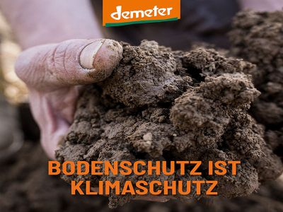 Auf dem Bild ist eine Hand zu sehen, die eine große Menge Erde hält. Über dem Motiv schwebt das Logo von Demeter. Unten am Bildrand steht in orangefarbener Schrift der Slogan "BODENSCHUTZ IST KLIMASCHUTZ"