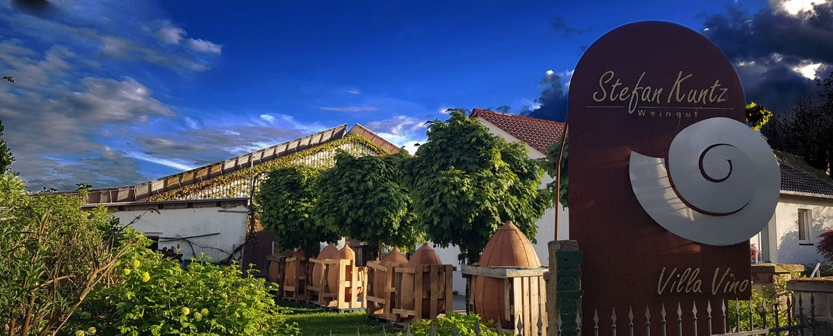  Ein rustikales Schild aus Cortenstahl mit der Aufschrift „Stefan Kuntz Weingut“ und „Villa Vino“ sowie einer stilisierten Schnecke steht im Vordergrund. Dahinter erstreckt sich ein Weingut oder ein ländliches Anwesen mit weißen Gebäuden und einem Dach, das von Weinreben bewachsen ist. Mehrere kleine, kugelförmig geschnittene Bäume säumen einen Weg. Im Hintergrund ist ein dramatischer Himmel mit blauen und dunkleren Wolken zu sehen.