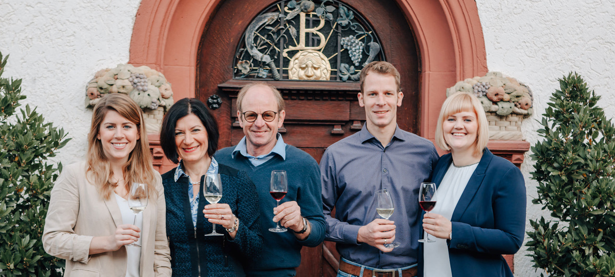  Fünf lächelnde Personen stehen vor einer aufwendig geschnitzten Holztür mit einem großen, verzierten "B" und Weinmotiven darüber. Die drei Frauen und zwei Männer halten alle Weingläser. Das Gebäude hat eine helle Fassade mit roten Akzenten und floralen Verzierungen über dem Türeingang. Grüne Sträucher oder Pflanzen säumen die Seiten des Eingangs.