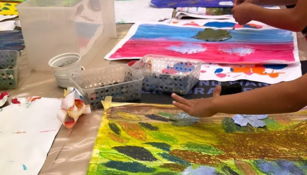 Eine Nahaufnahme von mehreren Kinderzeichnungen auf einem Tisch. Die Bilder sind mit Farbe gemalt und mit gestanzten Papierblumen verziert. Im Hintergrund sind Bastelmaterialien in Plastikboxen zu sehen. Eine Hand legt eine weitere Papierblume auf ein Bild.