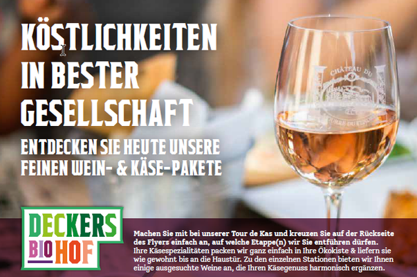 Ein Werbebild für Käse- und Weinpakete. Im Hintergrund ist ein gedeckter Tisch mit Käse zu sehen. Der Fokus liegt auf einem Glas Roséwein im Vordergrund auf der rechten Seite. Links ist in großen weißen Buchstaben der Text "KÖSTLICHKEITEN IN BESTER GESELLSCHAFT" zu lesen. Darunter steht in kleinerer Schrift "ENTDECKEN SIE HEUTE UNSERE FEINEN WEIN- & KÄSE-PAKETE". Am unteren Bildrand befindet sich das grüne Logo von "Deckers Biohof" und ein Text, der eine "Tour de Käse" bewirbt.
