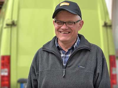 Mann mit Brille, grauer Jacke und Baseballcap, lächelt in die Kamera.