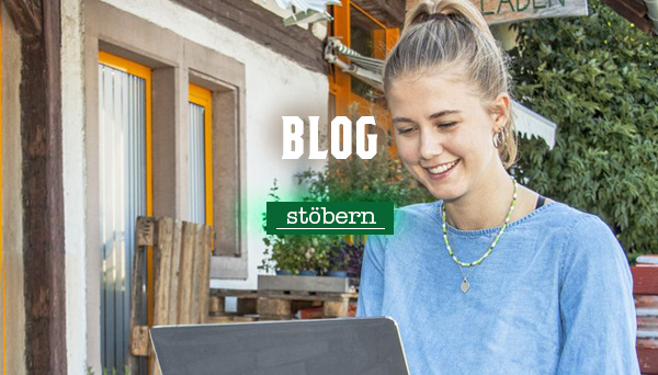 Junge Frau mit blondem Zopf sitzt draußen vor einem Laptop und lächelt. Im Hintergrund ein Haus mit Holzfassade. Text im Bild: „Blog – stöbern