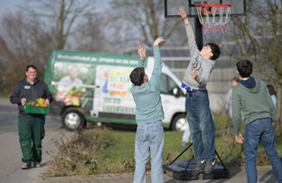 KI generiert: Kinder spielen Basketball, während ein Mann Kisten trägt. Ein Lieferwagen mit Werbung steht im Hintergrund.