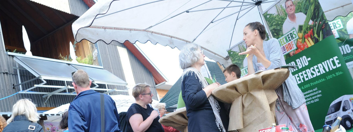 Menschen stehen unter einem großen weißen Sonnenschirm an einem mit Jutesack bedeckten Stehtisch auf einem Markt. Im Hintergrund Banner mit Werbung für einen Biolieferdienst „Beckers Biohof“. Eine Frau im Gespräch mit einer älteren Dame, weitere Besucher:innen im Austausch oder beim Schauen.