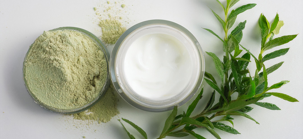  Ein flaches Glasgefäß mit hellgrünem Pulver steht neben einem offenen Tiegel mit weißer Creme auf einer weißen Oberfläche. Daneben liegen grüne Pflanzenzweige. Etwas von dem Pulver ist um das Glasgefäß herum verstreut.