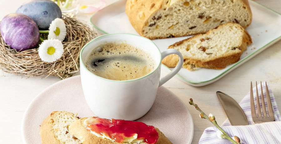 Eine frühlingshafte Frühstücksszene mit einer Tasse Kaffee, die auf einem rosafarbenen Teller steht. Daneben liegt ein aufgeschnittenes Brot mit Butter und roter Marmelade. Im Hintergrund sind auf einem weiteren Teller ein Stück Stollen oder Rosinenbrot und bunte Ostereier in einem Vogelnest zu sehen. Auf dem Tisch liegt ein Besteck-Set und ein Weidenkätzchenzweig.