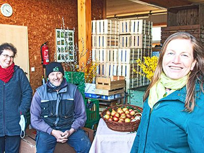 Zwei Frauen und ein Mann in einem Innenraum, in dem Äpfel sortiert und in Kisten gelagert werden.