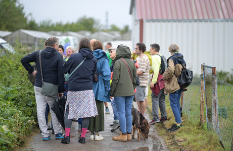 Gruppe von Menschen steht draußen zusammen, einige mit Rucksäcken und Hund, mit landwirtschaftlichen Gebäuden im Hintergrund.