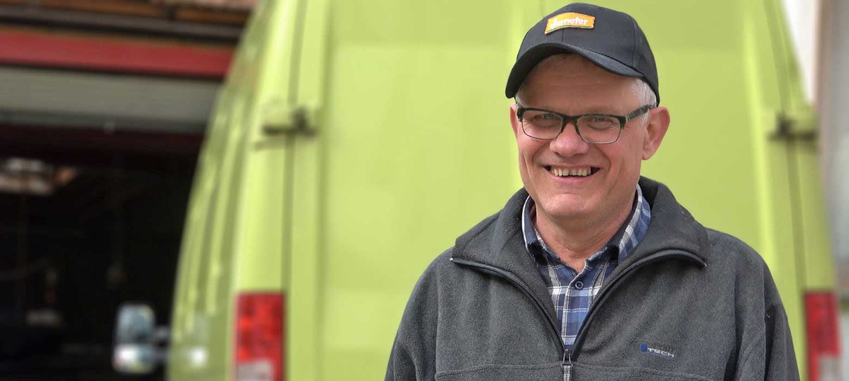  Ein lächelnder Mann mittleren Alters mit Brille, einer schwarzen Baseballkappe und einer grauen Fleecejacke steht vor der Rückseite eines hellgrünen Lieferwagens.