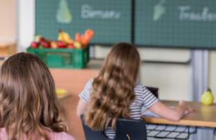 KI generiert: Zwei Kinder im Klassenzimmer mit Obstkiste. Text auf Tafel: "Bananen" und "Trauben".