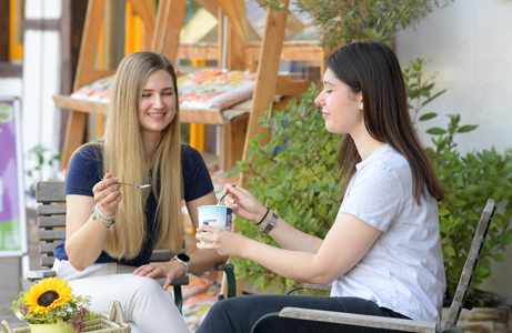 Zwei Frauen sitzen draußen an einem Tisch und essen gemeinsam Eis. Auf dem Tisch steht ein kleiner Korb mit Sonnenblumen.