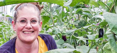 Ein Porträt einer lächelnden jungen Frau mit Brille. Sie trägt eine lila-gelbe Jacke und schaut zur Seite. Rechts von ihr sind große, grüne Pflanzen mit einer lila Aubergine zu sehen, die zwischen den Blättern hängt. Der Hintergrund ist unscharf, was auf ein Gewächshaus hindeutet.