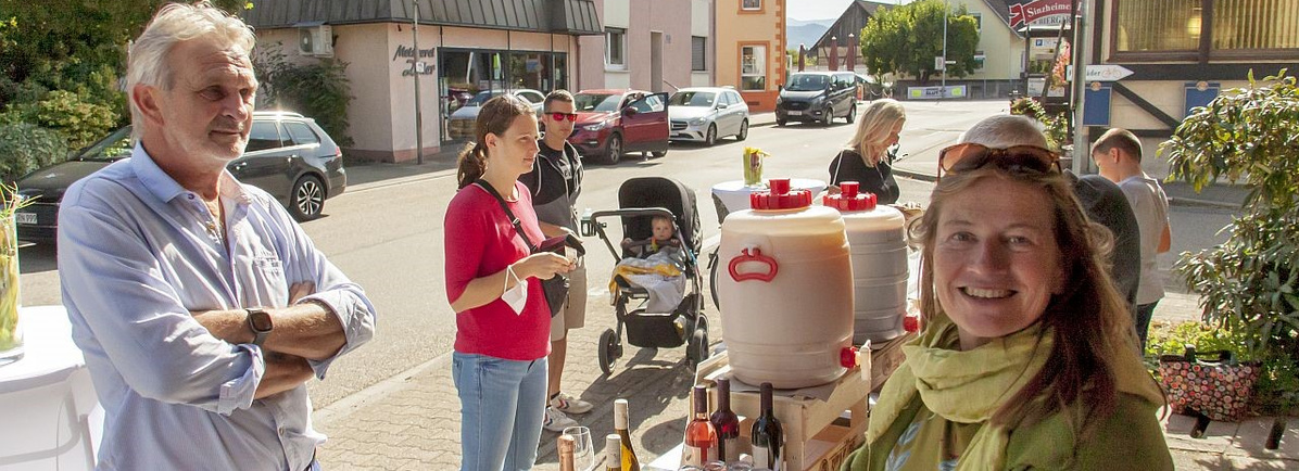 Eine Gruppe von Personen steht im Freien in einer Straße und unterhält sich. Im Vordergrund, rechts, lächelt Helga Decker, mit einem grünen Schal. Links steht ein Mann mit verschränkten Armen und einem ernsten Gesichtsausdruck. In der Mitte der Aufnahme, in der Nähe eines Kinderwagens, steht eine Frau mit einem Baby. Zwei große, weiße Behälter, möglicherweise für Getränke, stehen in der Mitte des Bildes. Im Hintergrund sind mehrere geparkte Autos zu sehen.