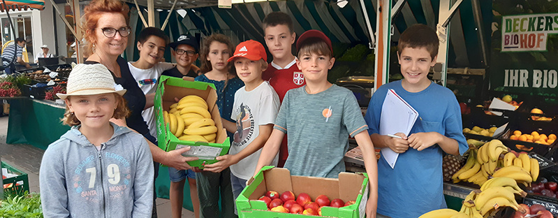 Eine Gruppe von sieben lachenden Kindern und einer Frau an einem Obststand. Zwei Jungen halten eine grüne Kiste mit Bananen, die sie gerade entgegennehmen. Im Hintergrund sind weitere Früchte und Marktstände zu sehen.
