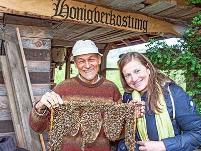 Zwei Personen zeigen eine große Wabe voller Bienen, darüber ein Schild ‚Honigverkostung‘.