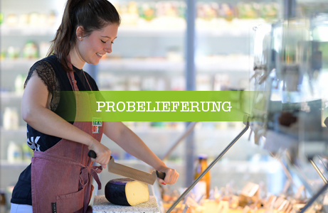Eine Mitarbeiterin schneidet an einer Käsetheke Käse, mit einem grünen Banner überlagert, auf dem 'PROBELIEFERUNG' steht.