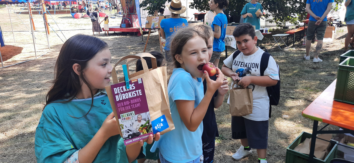 Eine Gruppe von Kindern steht an einem Tisch im Freien. Ein Mädchen mit langen braunen Haaren hält eine Papiertüte mit dem Aufdruck "Deckers Bürokiste". Ein anderes Mädchen in einem blauen T-Shirt isst einen roten Apfel. Ein Junge mit einem weißen Rucksack hält ebenfalls eine Papiertüte. Im Hintergrund sind weitere Tische und Bäume zu erkennen.
