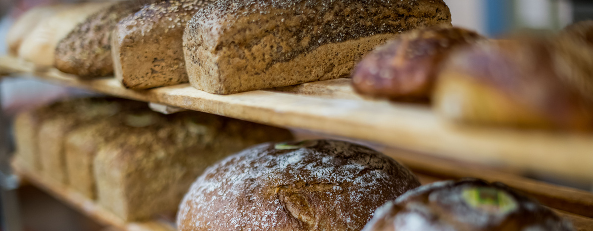 Eine Auswahl frisch gebackener Brote und Brotlaibe auf Holzregalen, einige davon mit Körnern oder Mehl bestäubt, in einem warmen, natürlichen Licht.