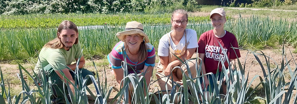 Eine Gruppe von vier lächelnden Personen – zwei Männer und zwei Frauen – kniet in einer Reihe von Lauchpflanzen auf einem Feld und blickt direkt in die Kamera. Im Hintergrund sind weitere Felder mit grünen Pflanzen unter einem blauen Himmel zu sehen.