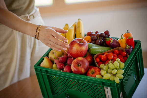 Geflochtener Korb mit frischem Obst, darunter Bananen, Äpfel, Birne, Zitrone und Trauben. Der Korb steht auf einem Holztisch.