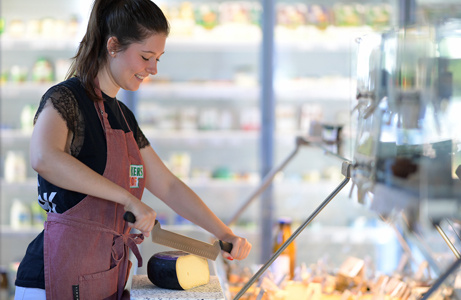 Eine Frau steht an der Käsetheke in einem Bioladen und schneidet ein großes Stück Käse. Im Hintergrund sind Regale mit weiteren Produkten sichtbar.
