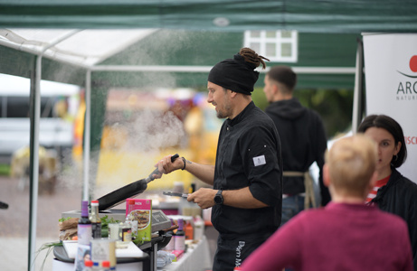 Ein Mann in schwarzer Kochjacke und Mütze kocht an einem Grill auf einem Stand im Freien, während eine andere Person zuschaut.