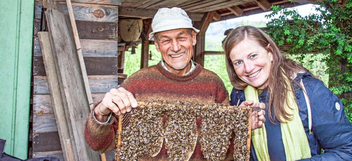  Ein lächelnder älterer Mann mit einem beigen Fischerhut und einem gestreiften Pullover hält zusammen mit einer lächelnden Frau in einer blauen Jacke und einem hellgrünen Schal einen Bienenstockrahmen voller Bienen. Sie stehen vor einem rustikalen Holzgebäude.