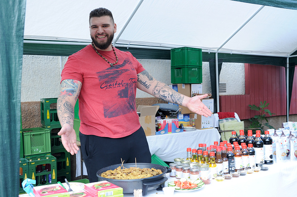 Ein Mann mit Tattoos und Bart in einem roten T-Shirt steht an einem Stand auf einem Hoffest und präsentiert eine Auswahl an Produkten.