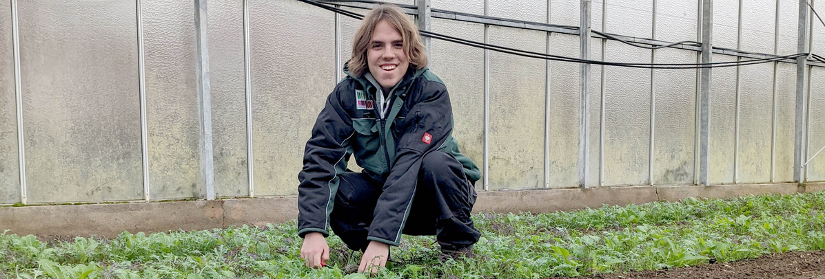 Ein junger Mann mit langen, blonden Haaren und einem Bart hockt in einem Gewächshaus zwischen Reihen junger Pflanzen. Er trägt eine dunkelgrüne Arbeitsjacke und eine schwarze Hose und lächelt in die Kamera.