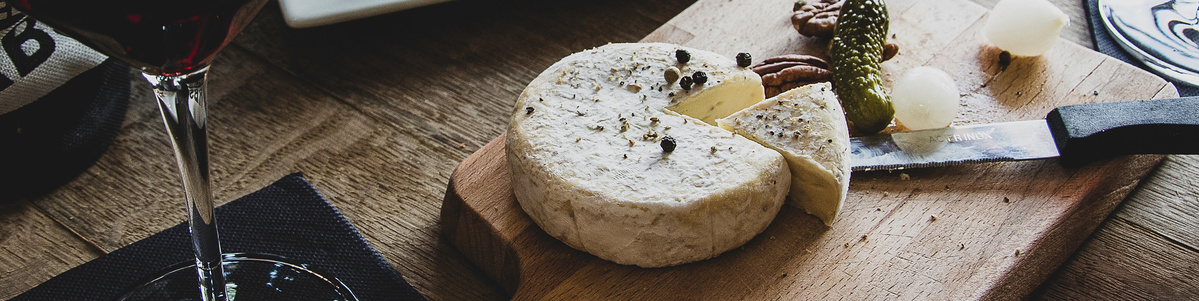 Ein aufgeschnittener Camembert mit Pfefferkörnern, daneben Perlzwiebeln, Cornichons und Nüsse auf einem Holzbrett. Dazu ein Glas Rotwein – serviert auf einem rustikalen Holztisch.