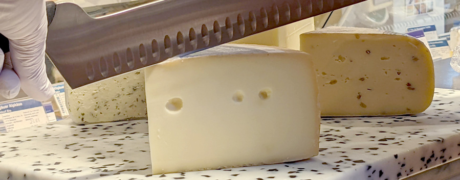 Eine Nahaufnahme von einer Hand mit einem weißen Handschuh, die ein großes, gezacktes Messer hält und damit in einen Laib Käse schneidet. Im Hintergrund sind weitere Käsesorten und Preisschilder zu sehen.