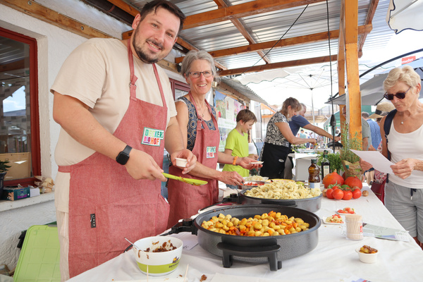 Ein lächelnder Mann in einer Kochschürze rührt in einer großen Pfanne auf einem Herd. Im Hintergrund sind weitere Personen zu sehen, die in einem überdachten Bereich sitzen oder stehen.