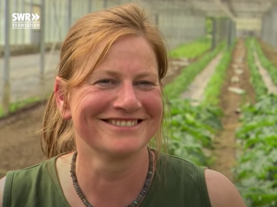 Screenshot aus einer SWR-Fernsehsendung: Nahaufnahme einer freundlich lächelnden Frau mit rötlich-blonden Haaren und einem grünen Oberteil. Sie steht in einem Gewächshaus, im unscharfen Hintergrund sind lange Reihen mit grünen Pflanzen zu sehen.