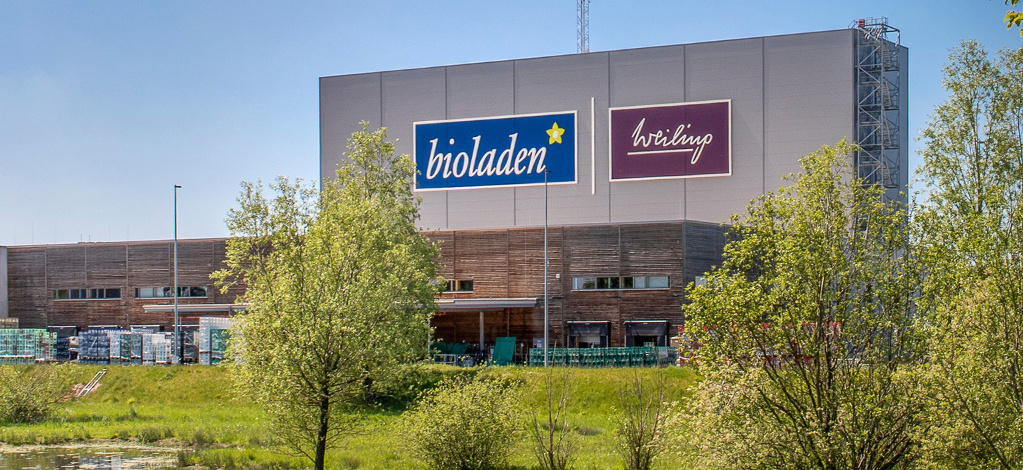  Ein modernes, großes Gebäude mit den Logos "bioladen" und "weiling" an der Fassade. Das Gebäude hat eine graue obere Hälfte und eine holzverkleidete untere Hälfte. Im Vordergrund sind grüne Bäume und Büsche zu sehen, und links unten ist ein kleines Gewässer oder eine feuchte Wiese angedeutet.