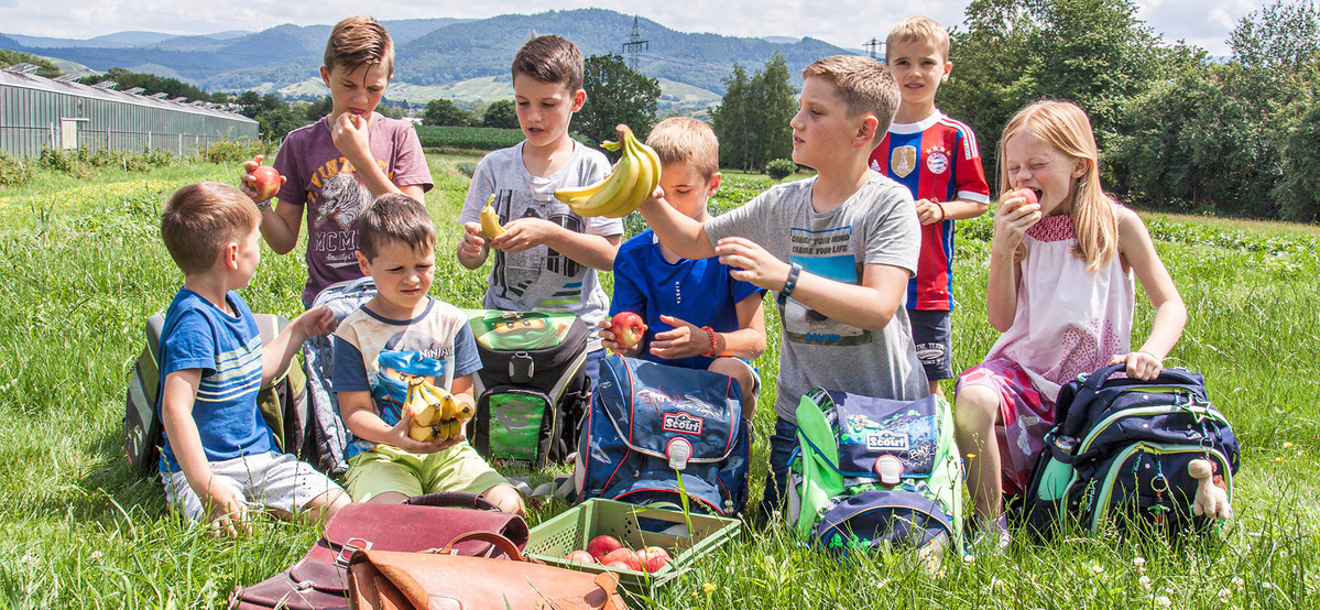 Acht Kinder genießen draußen nach der Schule einen gesunden Snack. Im Vordergrund erkennt man viele Schultaschen.