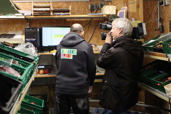Innenaufnahme: Zwei Männer stehen an einem Schreibtisch mit Computerbildschirm. Einer trägt eine Jacke mit dem Schriftzug ‚Deckers Biohof‘, ein Kameramann filmt die Szene.