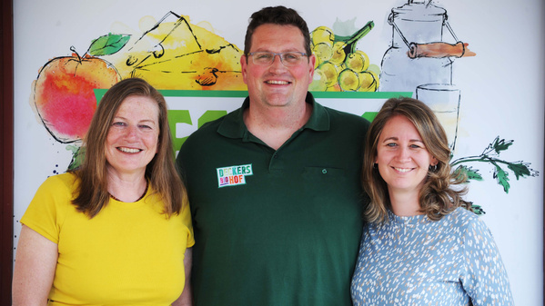 Drei Geschäftsführer stehen nebeneinander und lächeln in die Kamera. Hinter ihnen ist eine Wand mit gezeichneten Obst- und Gemüse-Motiven.